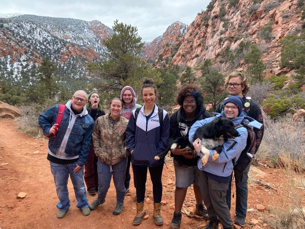 Group of smiling young adults and a dog on a snowy red-rock canyon trail during an Engage adventure outing